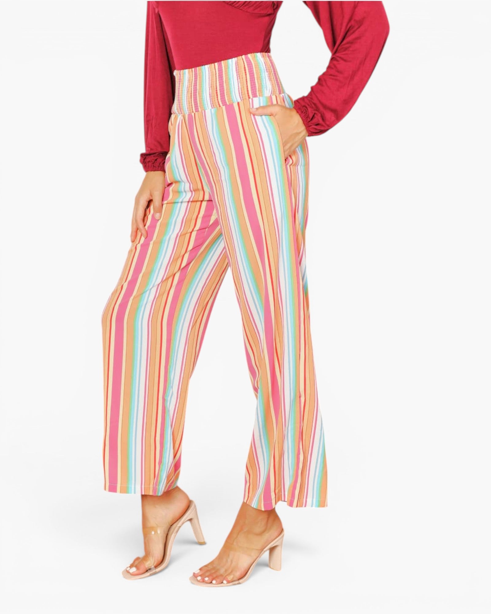 A model wearing high-waisted, wide-leg cropped pants with vertical multicolor stripes in pink, orange, yellow, green, and blue, styled with a red long-sleeve top and clear-strap high-heeled sandals against a white studio background.