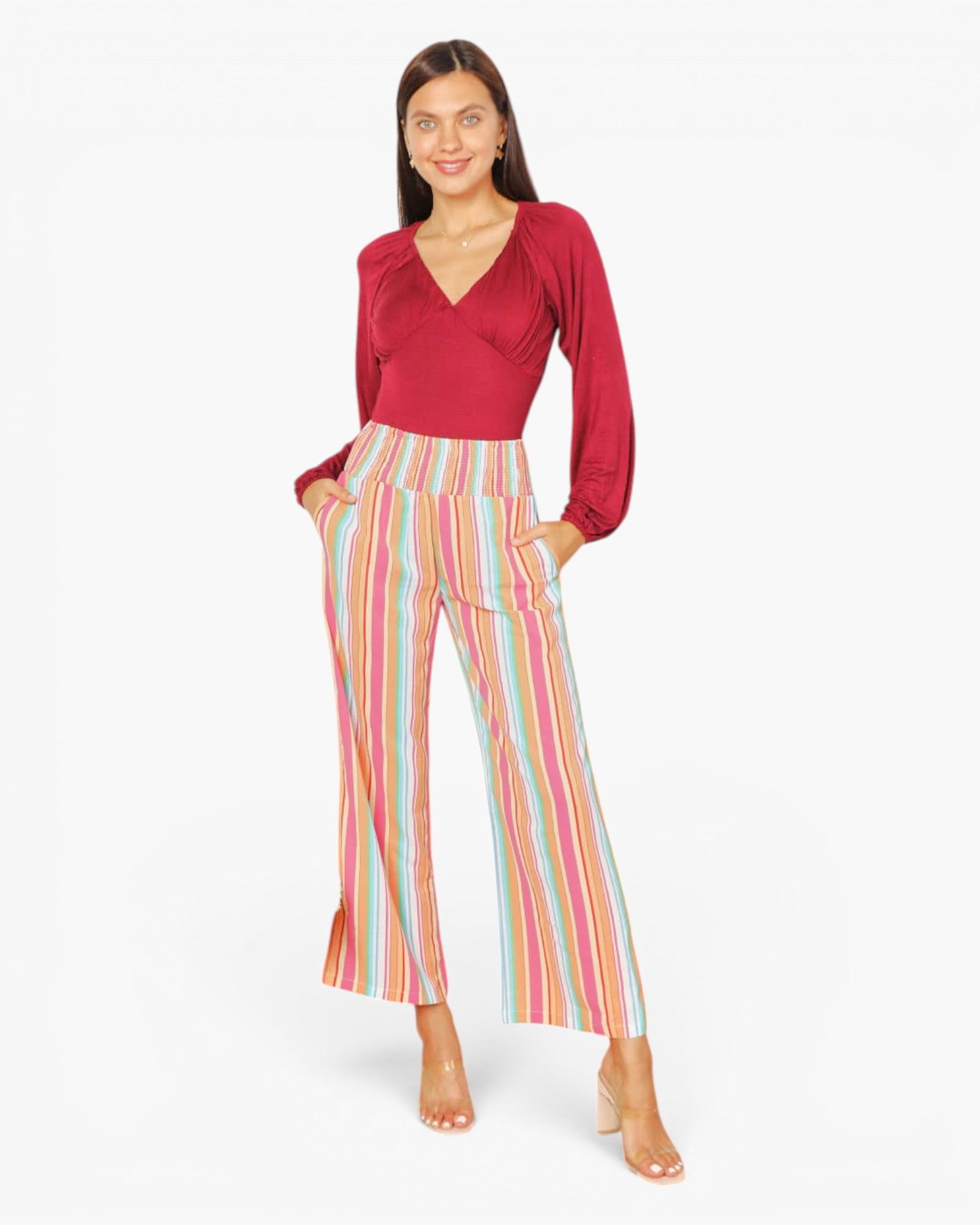A woman modeling a long-sleeve wrap-style top in deep red paired with high-waisted, wide-leg trousers featuring vertical pastel stripes in pink, yellow, blue, and green, set against a plain white studio background.