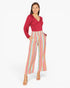 A woman modeling a long-sleeve wrap-style top in deep red paired with high-waisted, wide-leg trousers featuring vertical pastel stripes in pink, blue, yellow, and green, set against a plain white studio background.