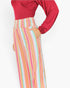 A model wearing a red long-sleeve top and high-waisted, wide-leg trousers with vertical pastel stripes in pink, orange, yellow, green, and blue, set against a plain white background.