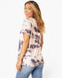 A woman wearing a short-sleeve tie-dye t-shirt in a light pink and dark gray pattern, paired with blue jeans, photographed against a solid white studio background.