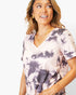 A woman wearing a short-sleeve V-neck tie-dye top in light pink and dark gray, paired with gold jewelry, against a plain white background.