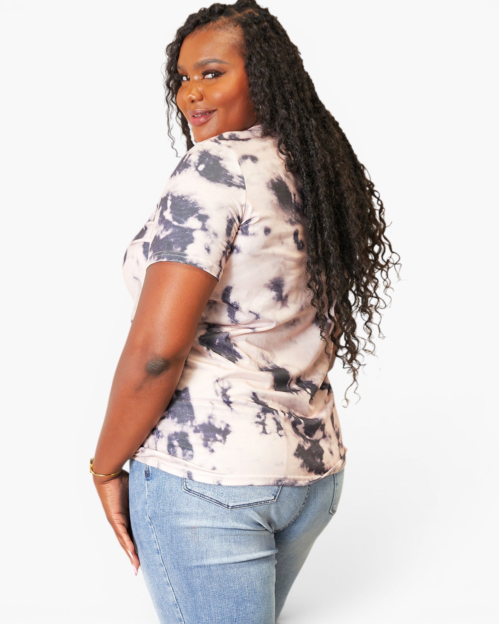 A woman with long, curly black hair is shown from a side-back angle wearing a short-sleeve tie-dye t-shirt with a beige and dark gray pattern, paired with light-wash blue jeans, against a plain white studio background.