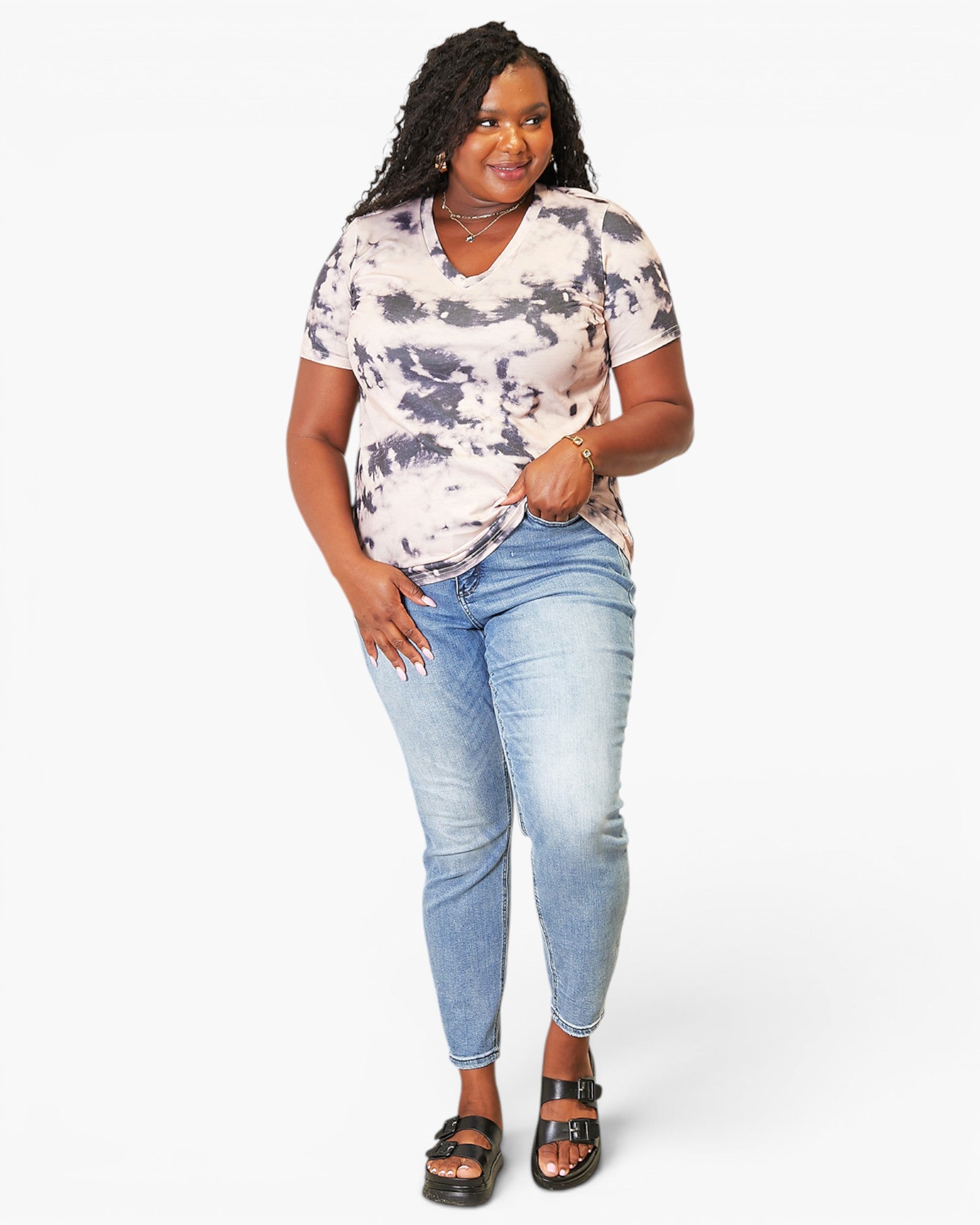 A woman modeling a short-sleeve V-neck tie-dye t-shirt in black and white, paired with light-wash blue jeans and black sandals, against a plain white studio background.