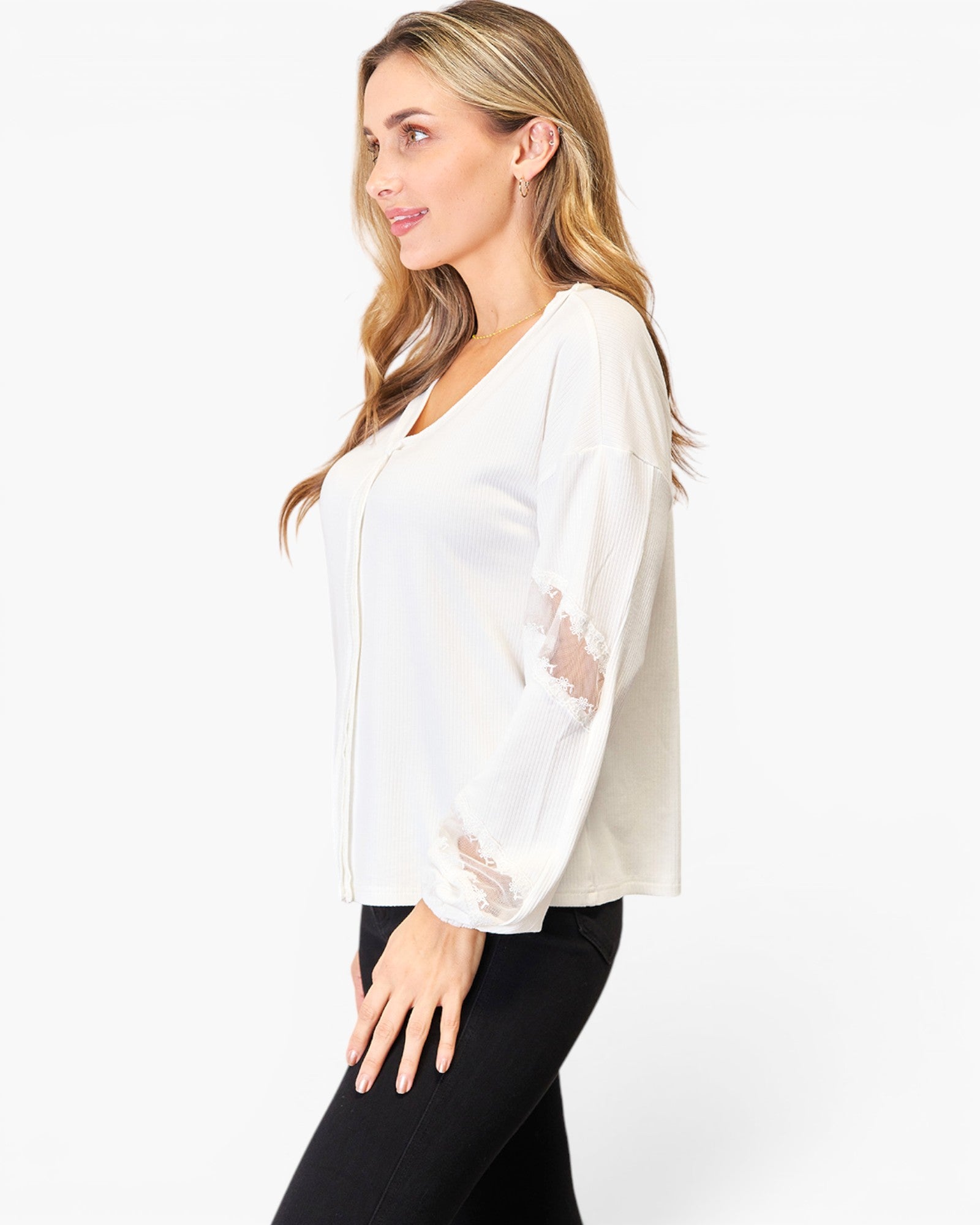A woman wearing a white long-sleeve blouse with sheer lace details on the sleeves, paired with black pants, photographed against a plain white studio background.