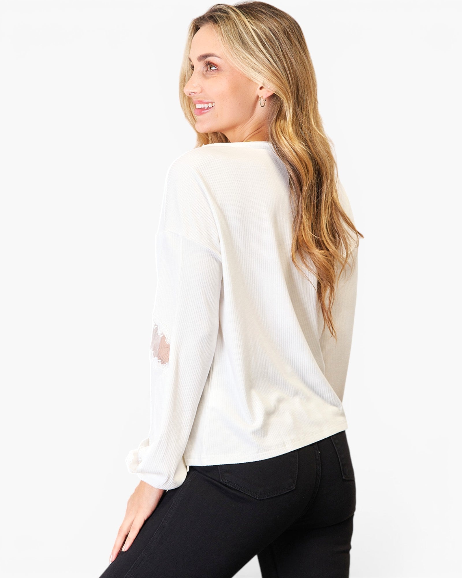 A woman with blonde hair is shown from a back-three-quarter angle wearing a cream-colored long-sleeve top with distressed details on the sleeves and black pants, set against a plain white studio background.