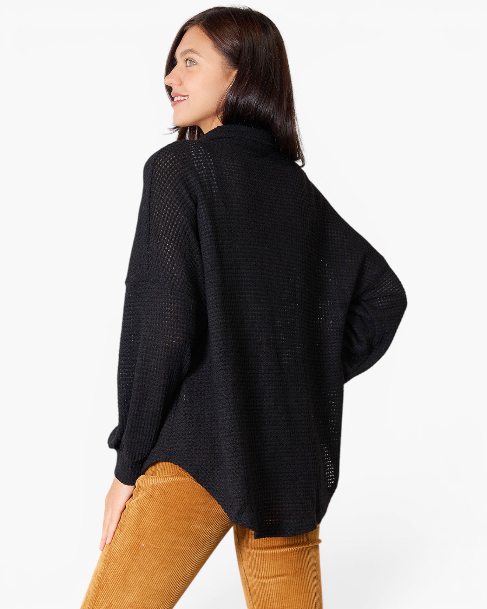 A woman wearing a black open-knit long-sleeve sweater and tan corduroy pants, photographed from the back against a plain white studio background.