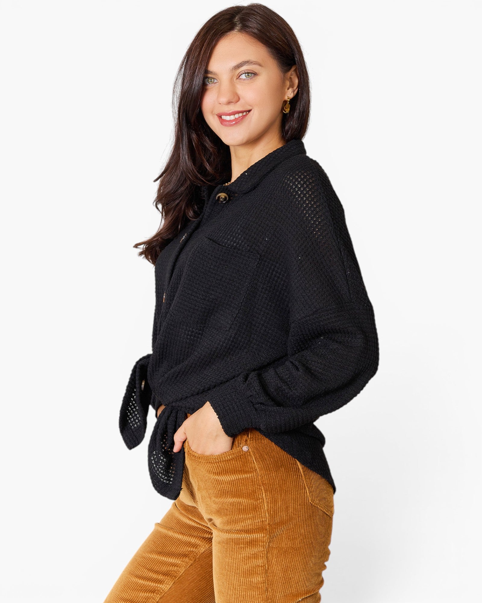 A woman wearing a black textured knit long-sleeve shirt with a tie waist and brown corduroy pants, photographed against a clean white background.