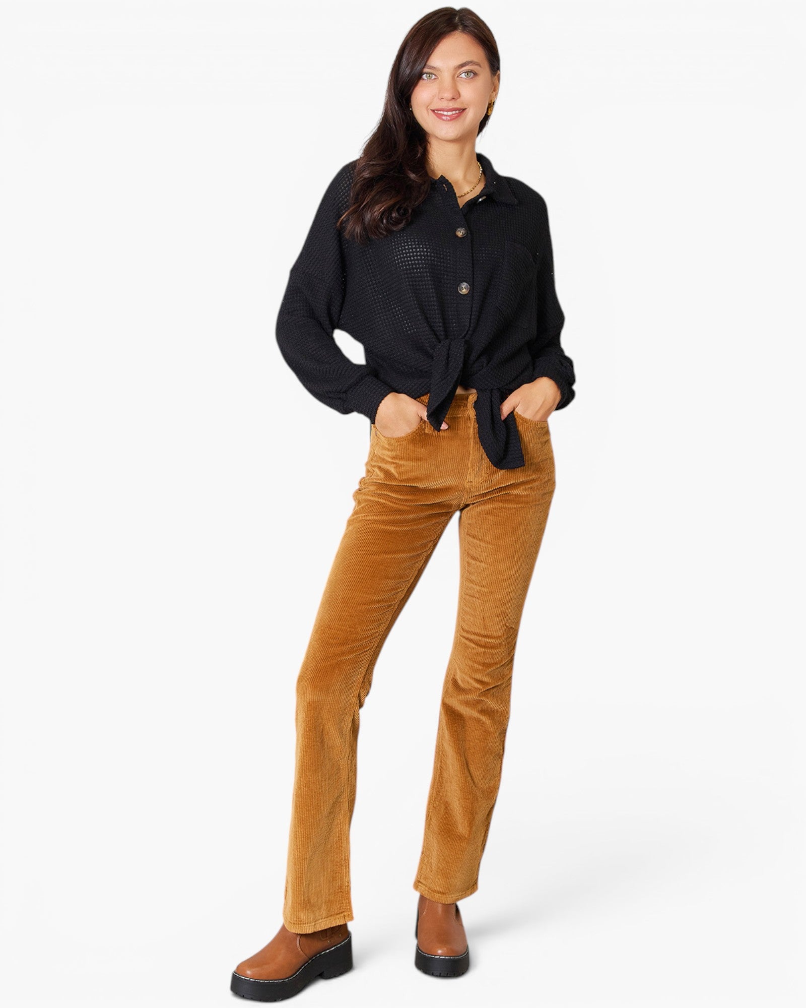 A woman modeling a black long-sleeve button-up shirt and brown corduroy pants against a light gray studio background.