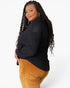 A woman with long black braids is shown from a side-back angle wearing a black textured knit sweater and mustard corduroy pants against a light gray studio background.