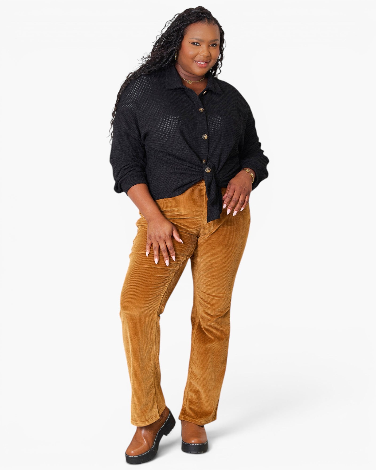 A woman modeling plus-size brown corduroy pants with a black button-down shirt and brown shoes, standing against a light gray studio background.