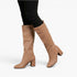 A pair of knee-high tan leather boots with a block heel, shown on a model wearing black pants against a light gray background.