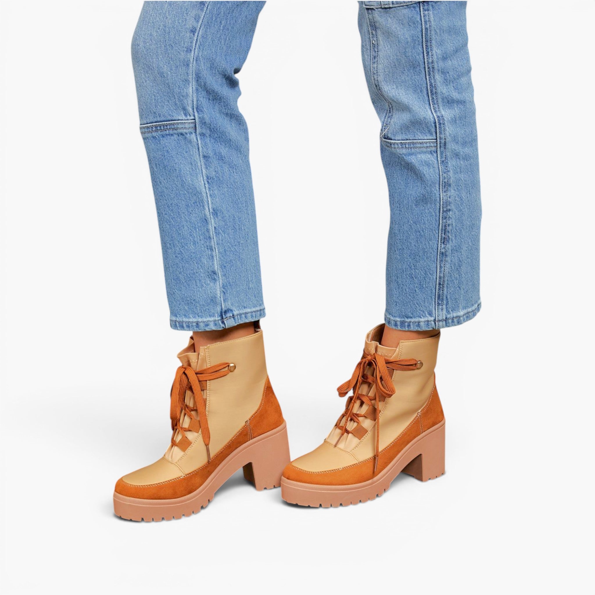 A person wearing light blue jeans and tan and brown lace-up ankle boots with chunky block heels, shown from the knees down on a light gray studio background.