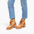 A person wearing light blue jeans and tan and brown lace-up ankle boots with chunky block heels, shown from the knees down on a light gray studio background.
