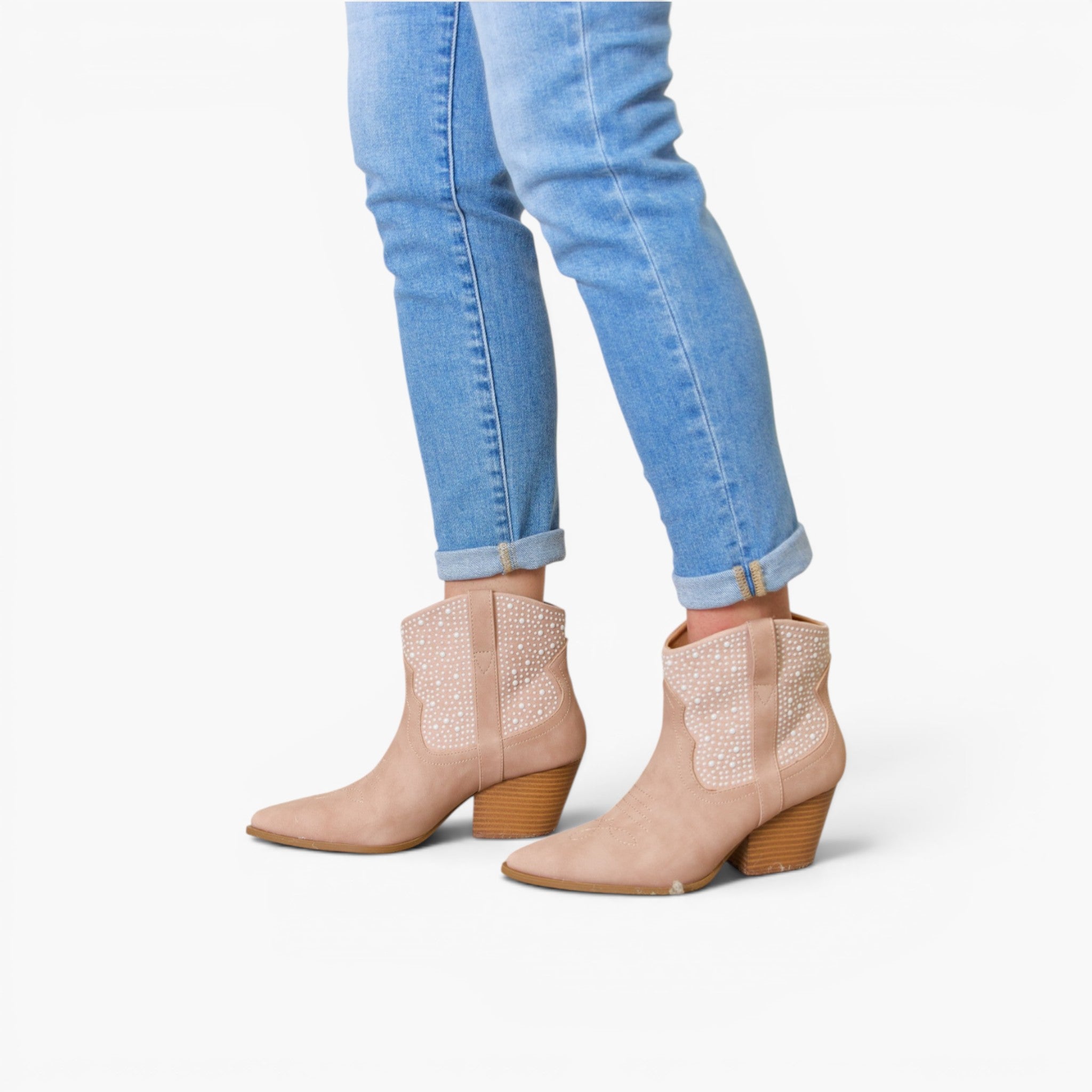 A person wearing light beige pointed-toe ankle boots with textured side panels and brown block heels, paired with light blue cuffed jeans, against a plain white background.