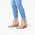 A person wearing light beige pointed-toe ankle boots with textured side panels and brown block heels, paired with light blue cuffed jeans, against a plain white background.