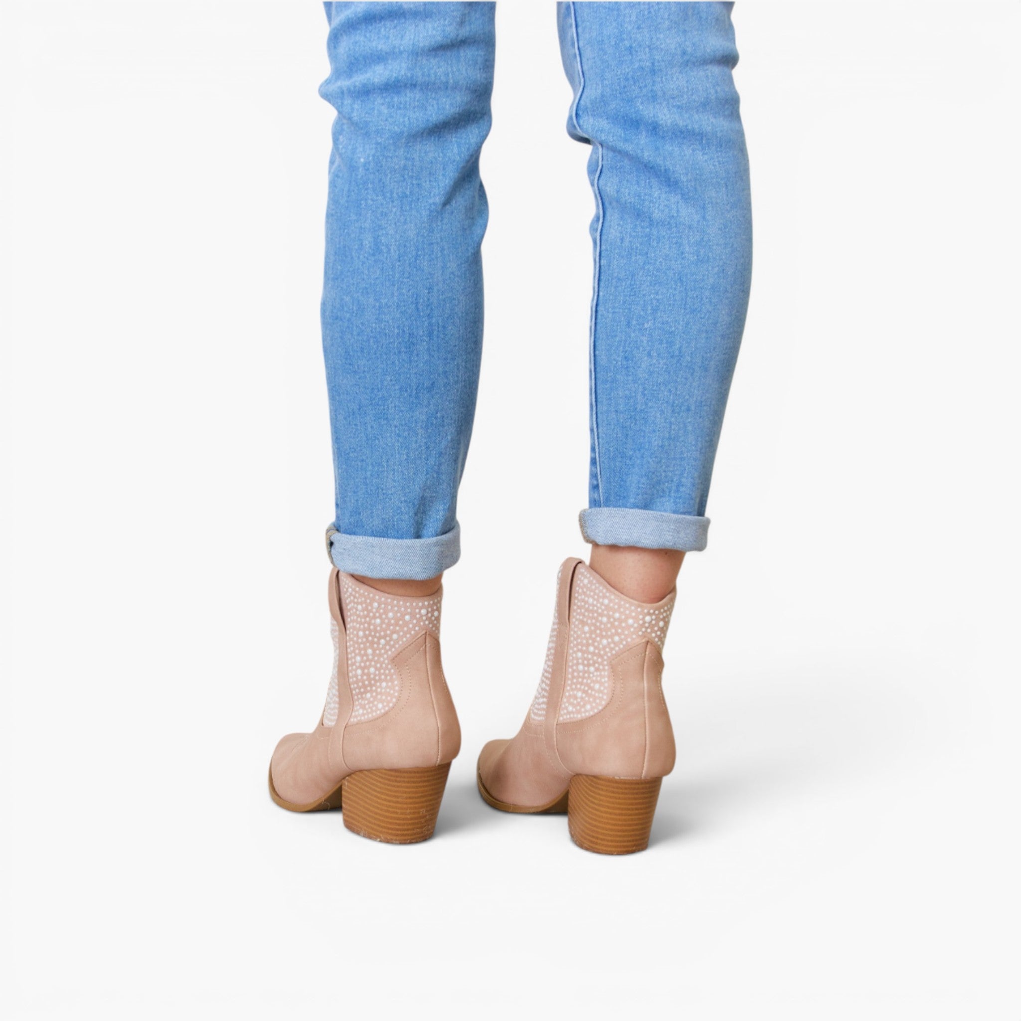 Rear view of a person wearing light beige ankle boots with textured side panels and brown block heels, paired with light blue cuffed jeans on a white background.