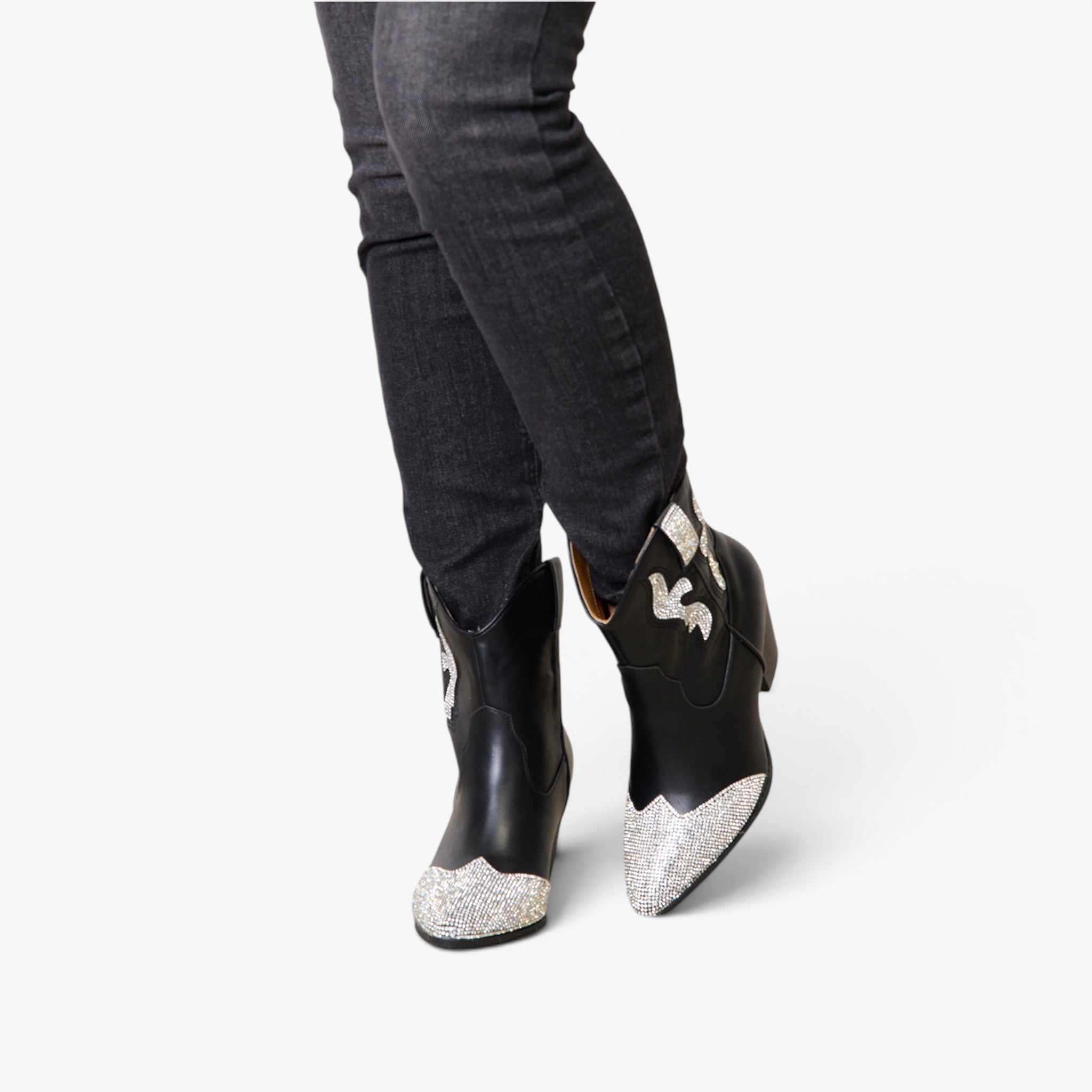 Black leather cowboy boots with silver glitter toe caps and white embroidered details, shown on a model wearing dark gray jeans against a white background.