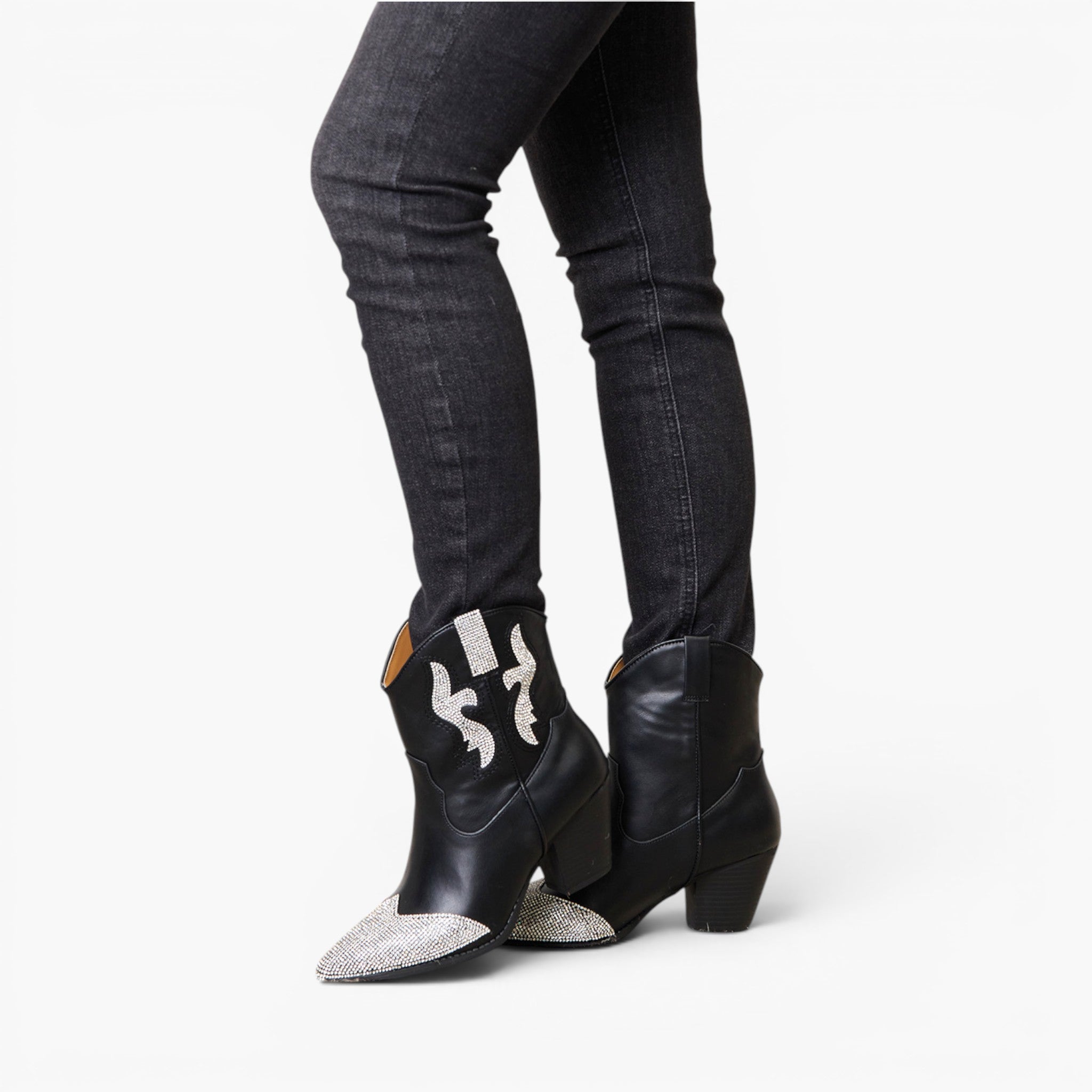 Black leather cowboy boots with silver glitter pointed toes and white embroidered details on the shaft, shown on a model wearing dark gray skinny jeans against a white background.