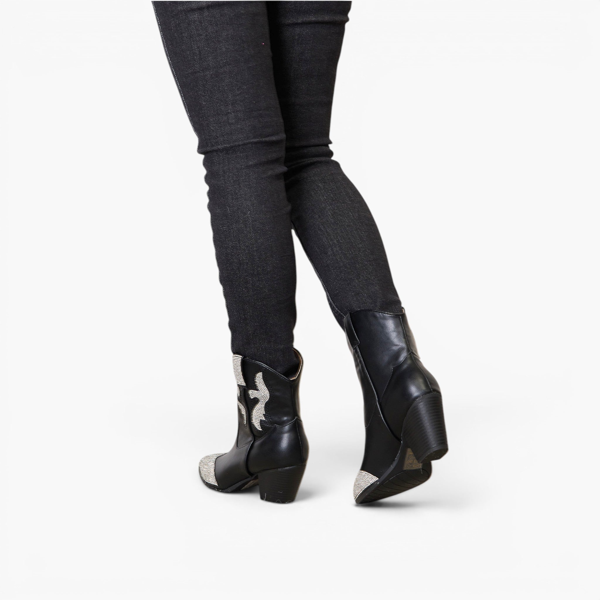 Black leather ankle boots with silver-toned western-style stitching and toe caps, shown on a model wearing dark gray skinny jeans from a rear three-quarter angle against a white studio background.