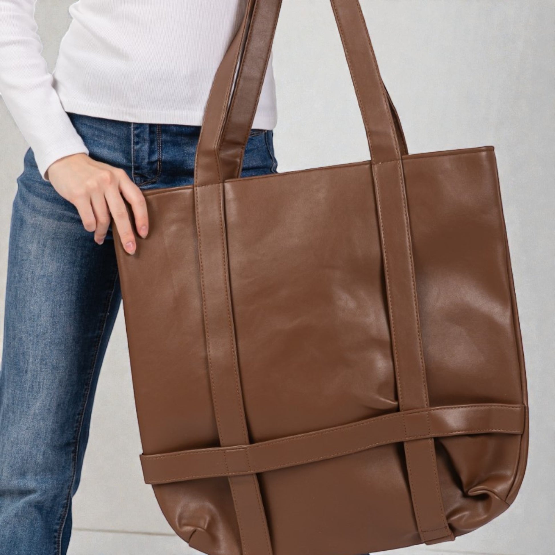 Brown leather tote bag held by a person wearing jeans and a white shirt on a light gray background