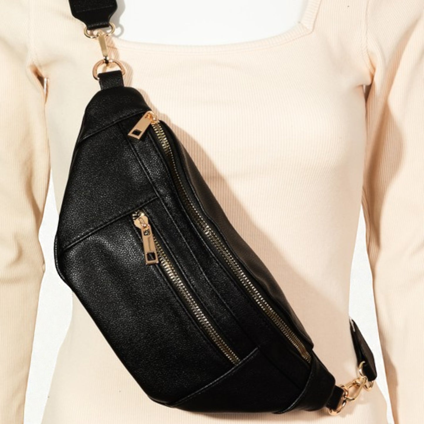 Black leather sling bag with gold zippers worn by a person in a beige outfit.