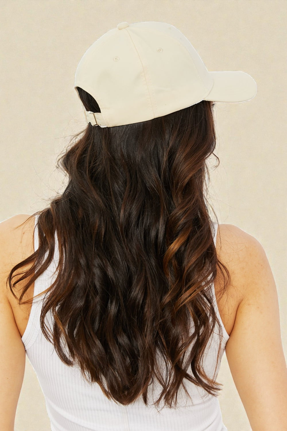 Person wearing a beige cap and white tank top with long, wavy hair on a beige background
