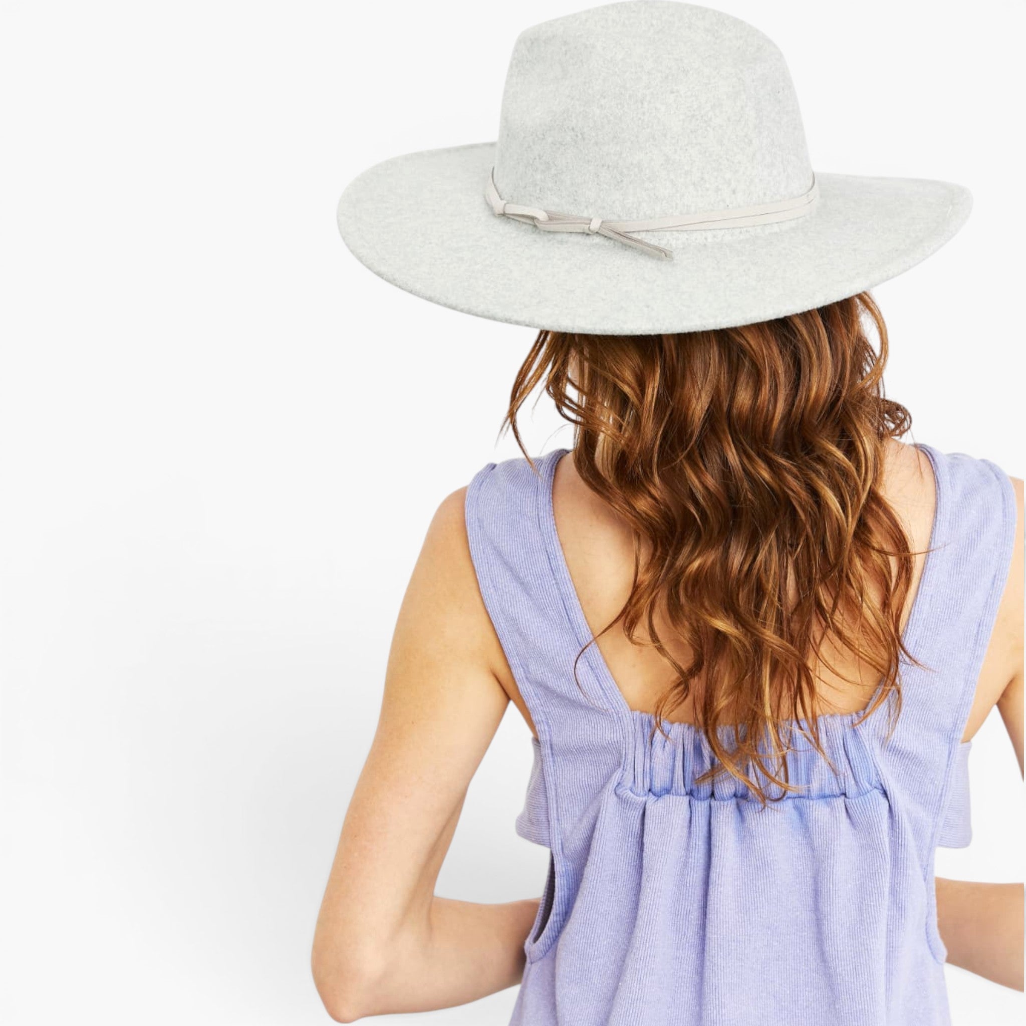 A woman shown from behind wearing a light gray felt wide-brim hat with a thin ribbon band and bow, paired with a lavender sleeveless top against a plain white background.