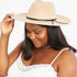 A woman wearing a beige wide-brim fedora hat with a black ribbon band, holding the brim with one hand against a plain white background.