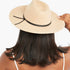 A woman viewed from behind wearing a beige wide-brim fedora hat with a black ribbon band, against a plain white background.