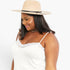 A woman wearing a beige wide-brim fedora hat with a black band and a white camisole top with lace trim, photographed against a plain light background.