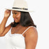 A woman wearing a light beige wide-brim fedora hat with a black band and a small gold buckle, paired with a white camisole top, photographed against a plain white background.