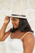 Woman wearing a beige hat with a black band against a textured beige wall.