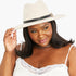 A woman wearing a light beige wide-brim fedora hat with a black band and a small gold triangle pin, paired with a white lace-trim camisole against a plain white background.