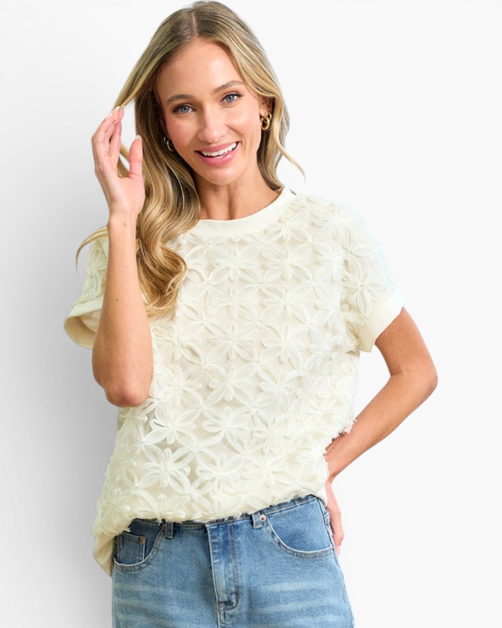 A woman modeling a cream-colored short-sleeve knit top with a textured, raised floral star pattern, paired with blue jeans against a plain white background.