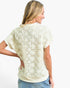 A woman shown from the back wearing a cream-colored short-sleeve top with a textured, raised floral starburst pattern, paired with light blue jeans against a plain white background.