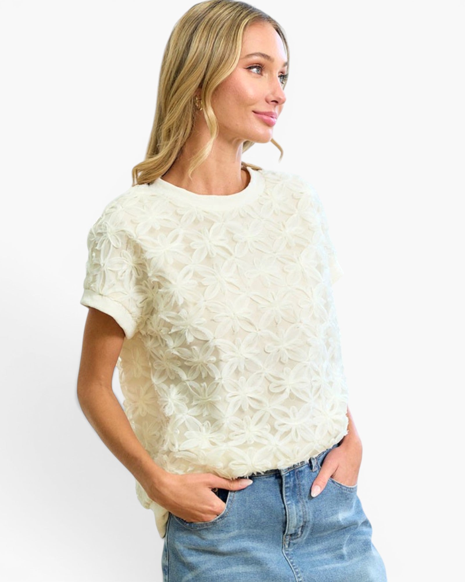 A woman wearing a cream-colored short-sleeve knit top with a textured, raised floral starburst pattern, paired with blue jeans, against a plain white background.