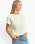 A woman wearing a cream-colored short-sleeve knit top with a textured, raised floral starburst pattern, paired with blue jeans, against a plain white background.