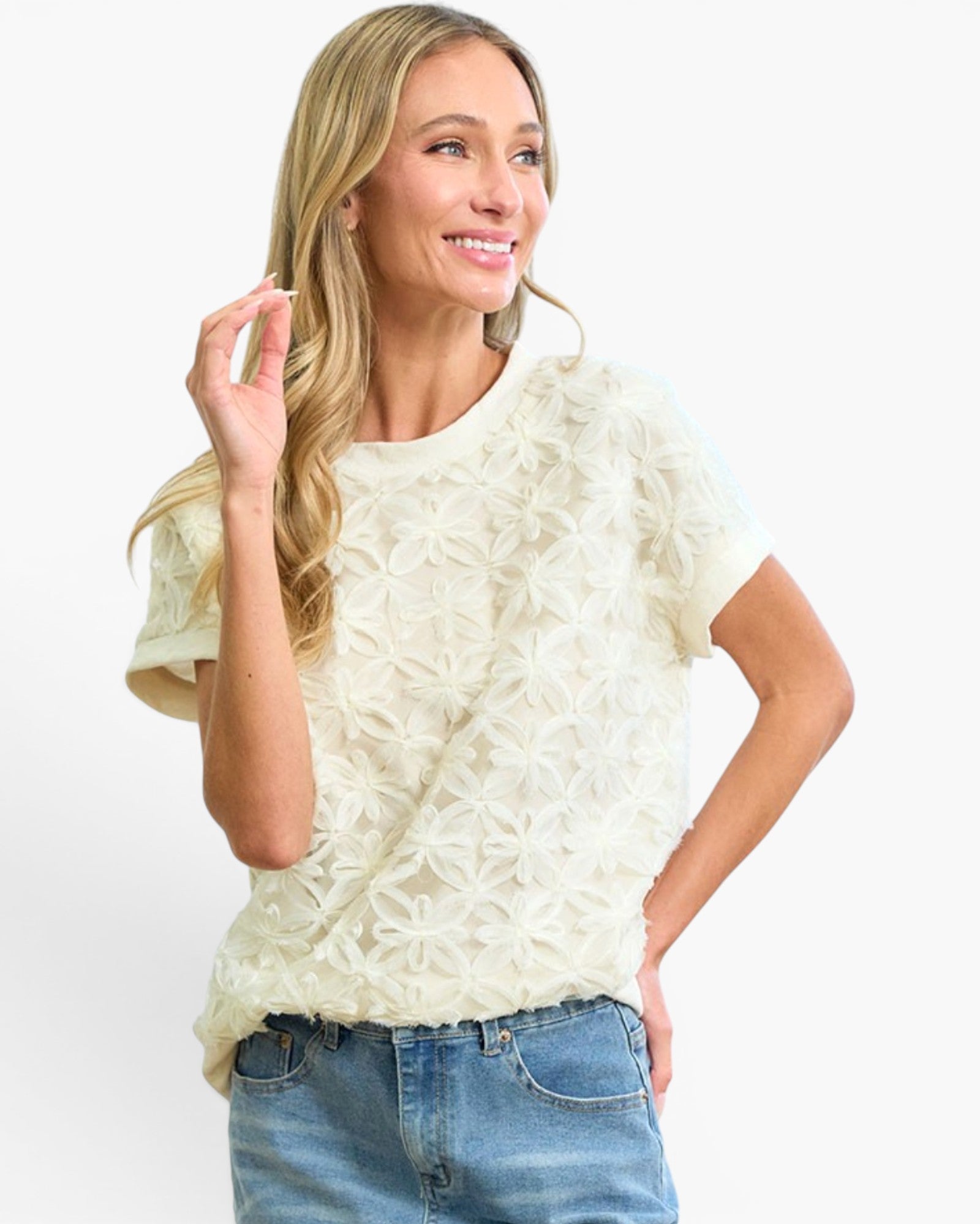 A woman wearing a cream-colored short-sleeve knit top with a textured, raised floral pattern, paired with blue jeans, against a plain white studio background.