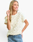 A woman wearing a cream-colored short-sleeve knit top with a textured, raised floral pattern, paired with blue jeans, against a plain white studio background.
