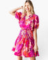 A woman modeling a short-sleeve mini dress with a vibrant pink, orange, and purple floral print against a plain white background.