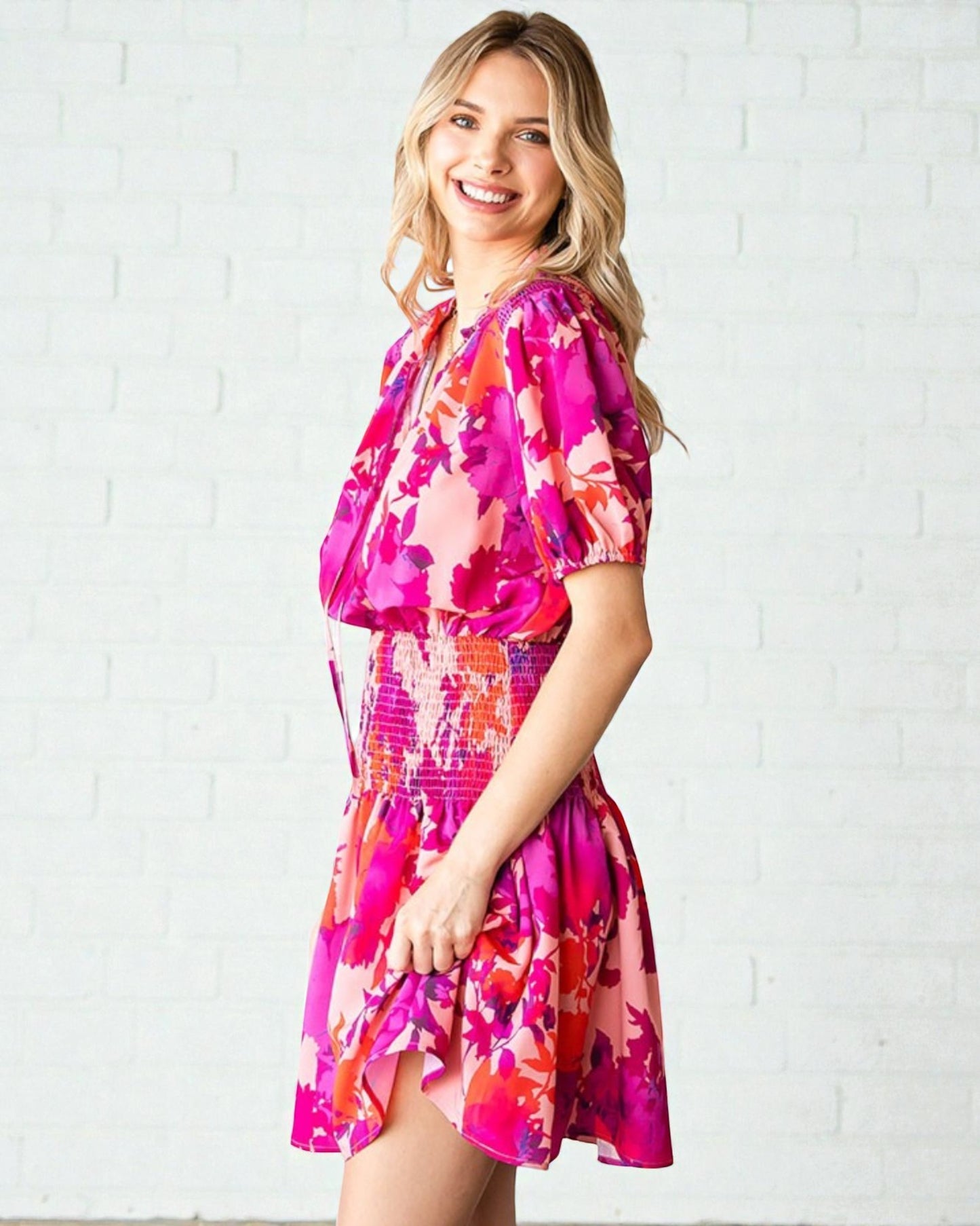 Woman wearing a vibrant pink floral dress against a white brick wall.