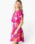 A woman modeling a short puff-sleeve dress with a vibrant abstract floral print in pink, magenta, orange, and white, set against a plain white studio background.