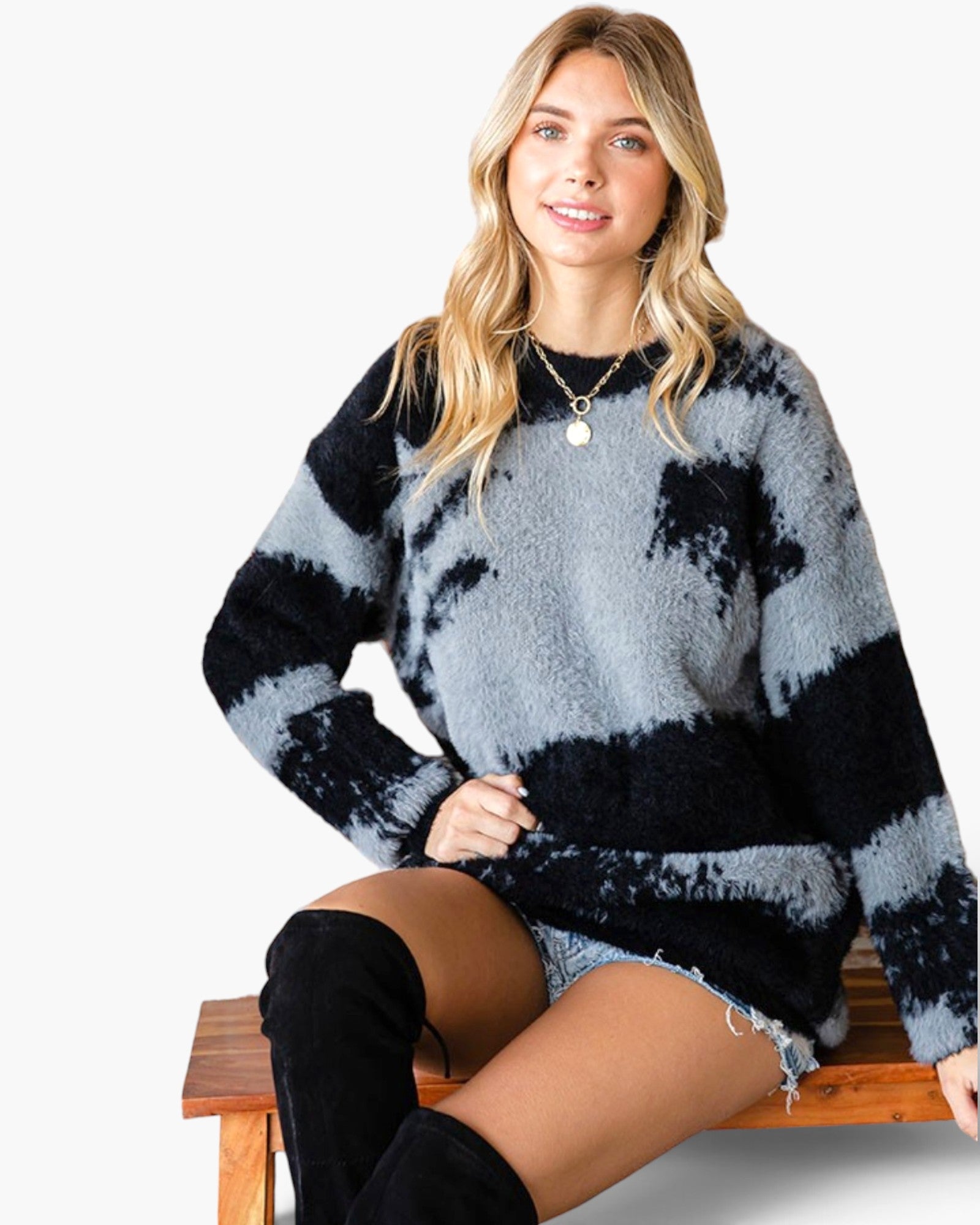 A woman wearing a fuzzy knit sweater with a black and light gray tie-dye pattern, sitting on a wooden bench against a plain white background.