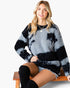 A woman wearing a fuzzy knit sweater with a black and light gray tie-dye pattern, sitting on a wooden bench against a plain white background.