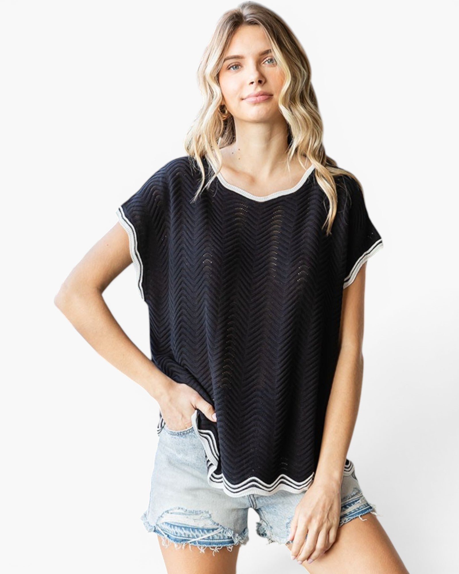 A woman modeling a black short-sleeve top with a subtle chevron pattern and white trim, paired with light-wash denim shorts, against a plain white background.