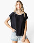 A woman modeling a black short-sleeve top with a subtle chevron pattern and white trim, paired with light-wash denim shorts, against a plain white background.