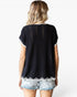 A woman shown from the back wearing a black short-sleeve knit top with white contrast trim and light-wash denim shorts against a plain white studio background.