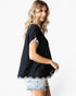 A woman wearing a black short-sleeve top with white contrast trim and a scalloped hem, paired with light-wash denim shorts, photographed against a plain white studio background.