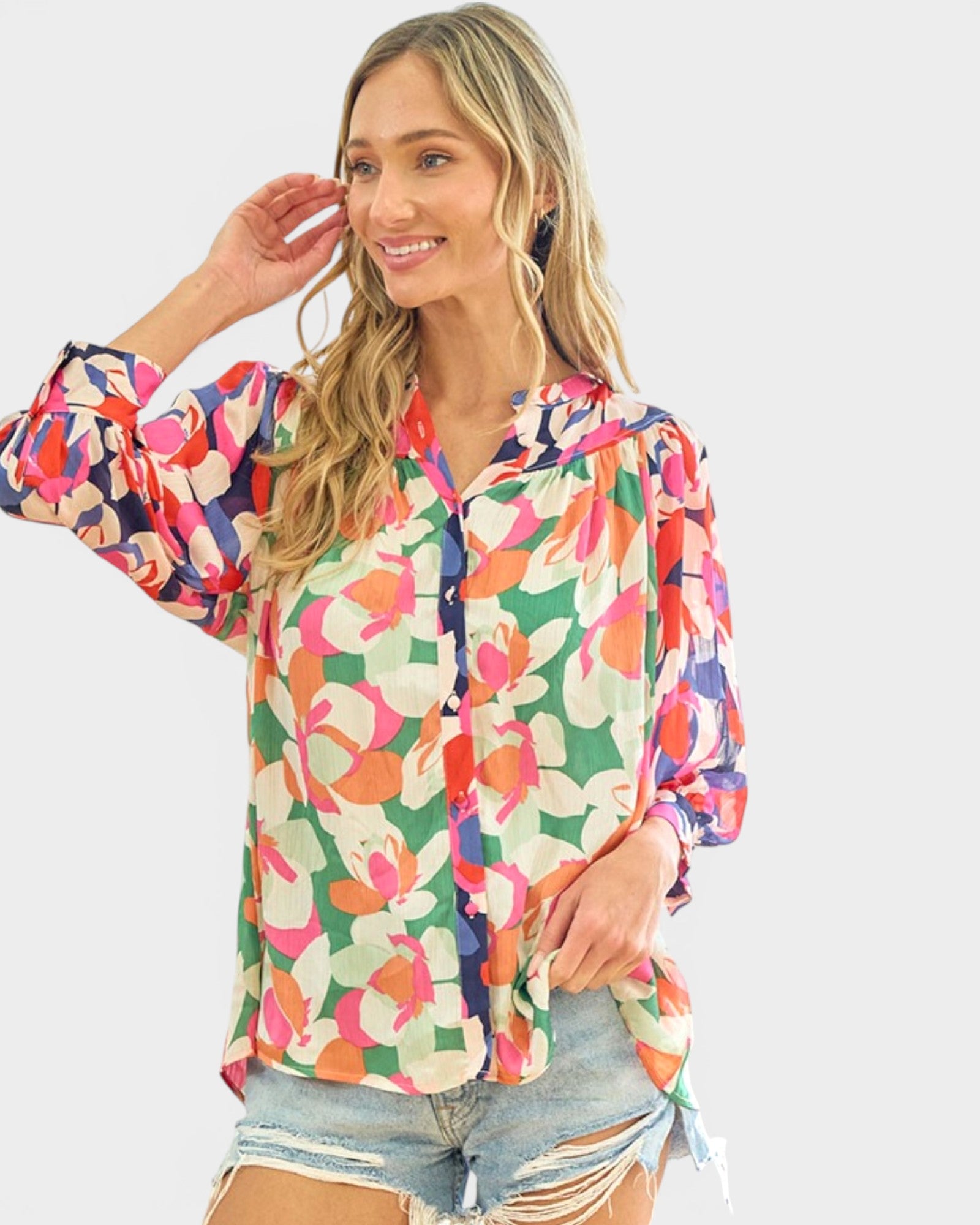 A woman wearing a colorful floral print button-up blouse with sheer puff sleeves and light-wash distressed denim shorts, posed against a solid light gray background.